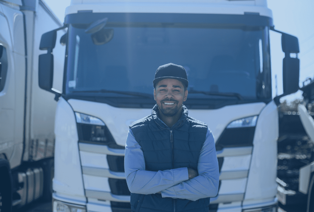 Young man in a baseball cap with arms folded standing infront of an HGV truck, smiling