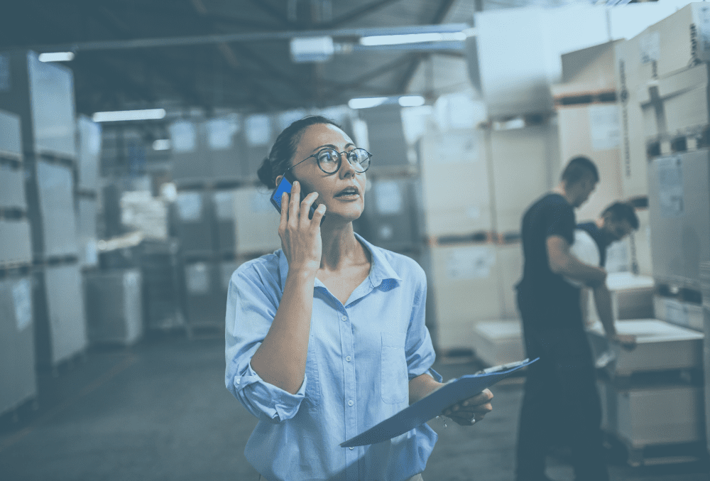 a female operations manager in a warehouse setting speaks on the phone and carries a clipboard