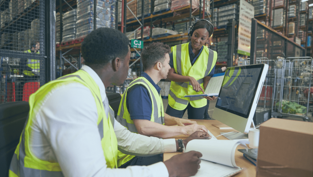 A woman in a hi-vis yellow jacket inside a warehouse talking to two men sitting down at a desk, also wearing hi-vis jackets