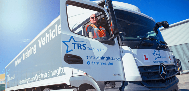 Angled view of an HGV Truck with TRS Training Vehicle written on the side and the cab door open with a man in hi-vis jacket standing up on the cab step, smiling, against bright blue sky