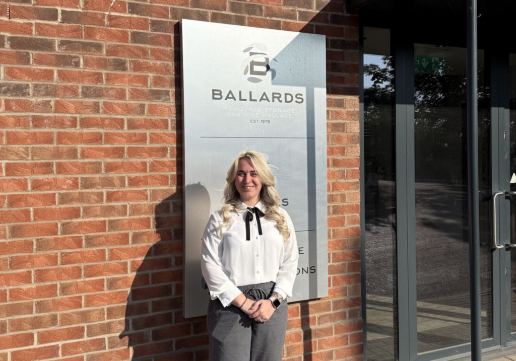 Abigail smiles while standing in front of a brick wall and a silver plaque displaying the Ballards Removals Ltd logo.
