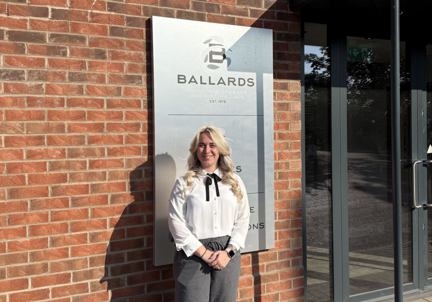 Abigail smiles while standing in front of a brick wall and a silver plaque displaying the Ballards Removals Ltd logo.