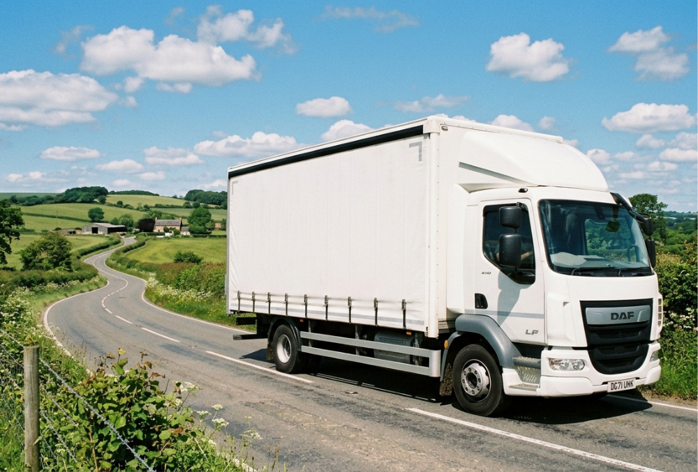A plain white Category C rigid-sided lorry driving along a winding country road, surrounded by green fields, hedgerows, and a blue sky with light white clouds.