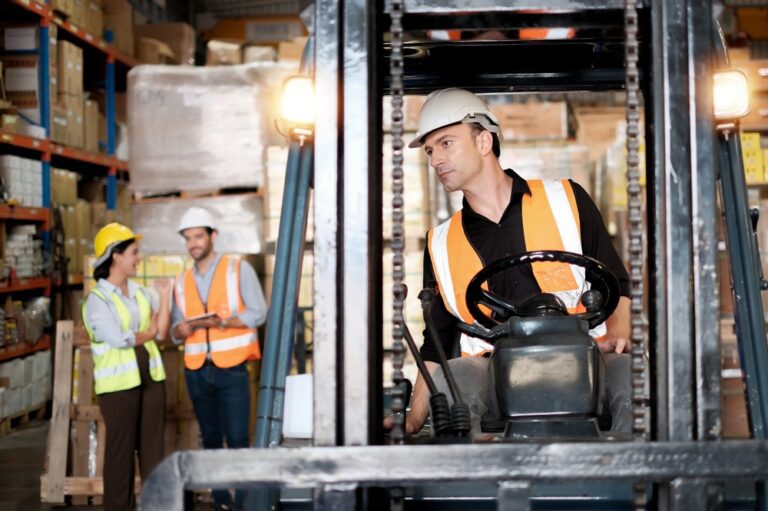A man in a hard hat drives a forklift truck in a warehouse with two colleagues standing talking in the background