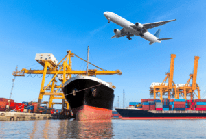 a big cargo ship in a port in front of cranes and an aeroplane in the air flying above it